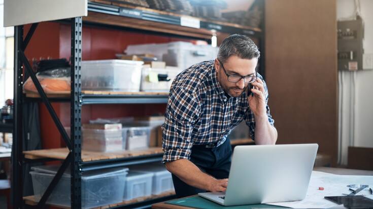 Shot of a man using a laptop and mobile phone while working on a project in a workshop