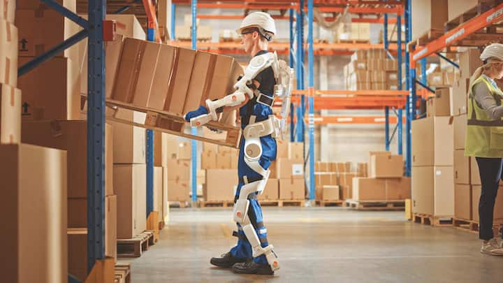 High-Tech Futuristic Warehouse: Worker Wearing Advanced Full Body Powered exoskeleton, Lifts Heavy Pallet full of Cardboard Boxes. Delivery Exosuit amplifies strength.
